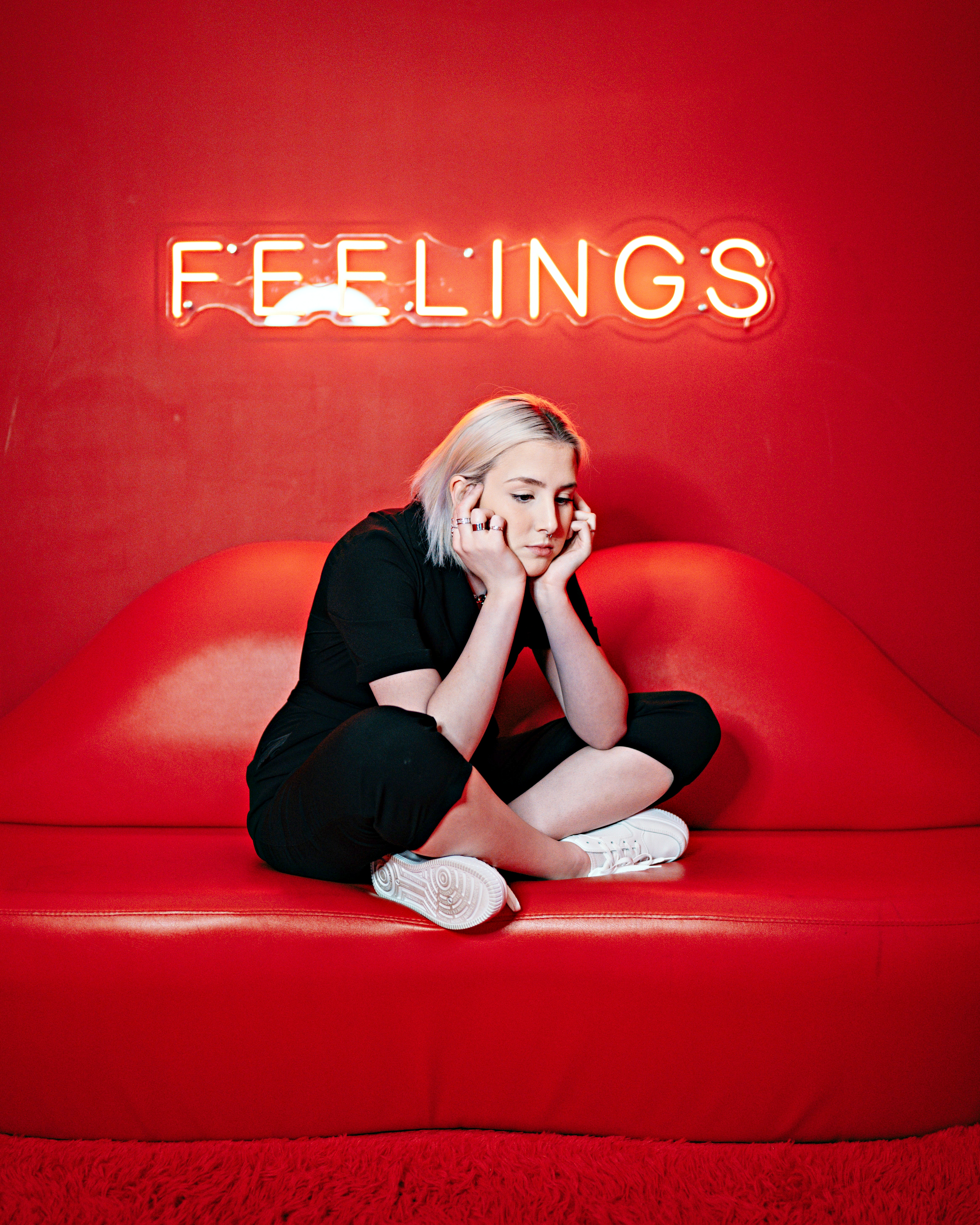 Gefühle-Liste – junge Frau mit blonden kinnlangen Haaren sitzt im Schneidersitz vor rotem Hintergrund mit der Aufschrift "FEELINGS"