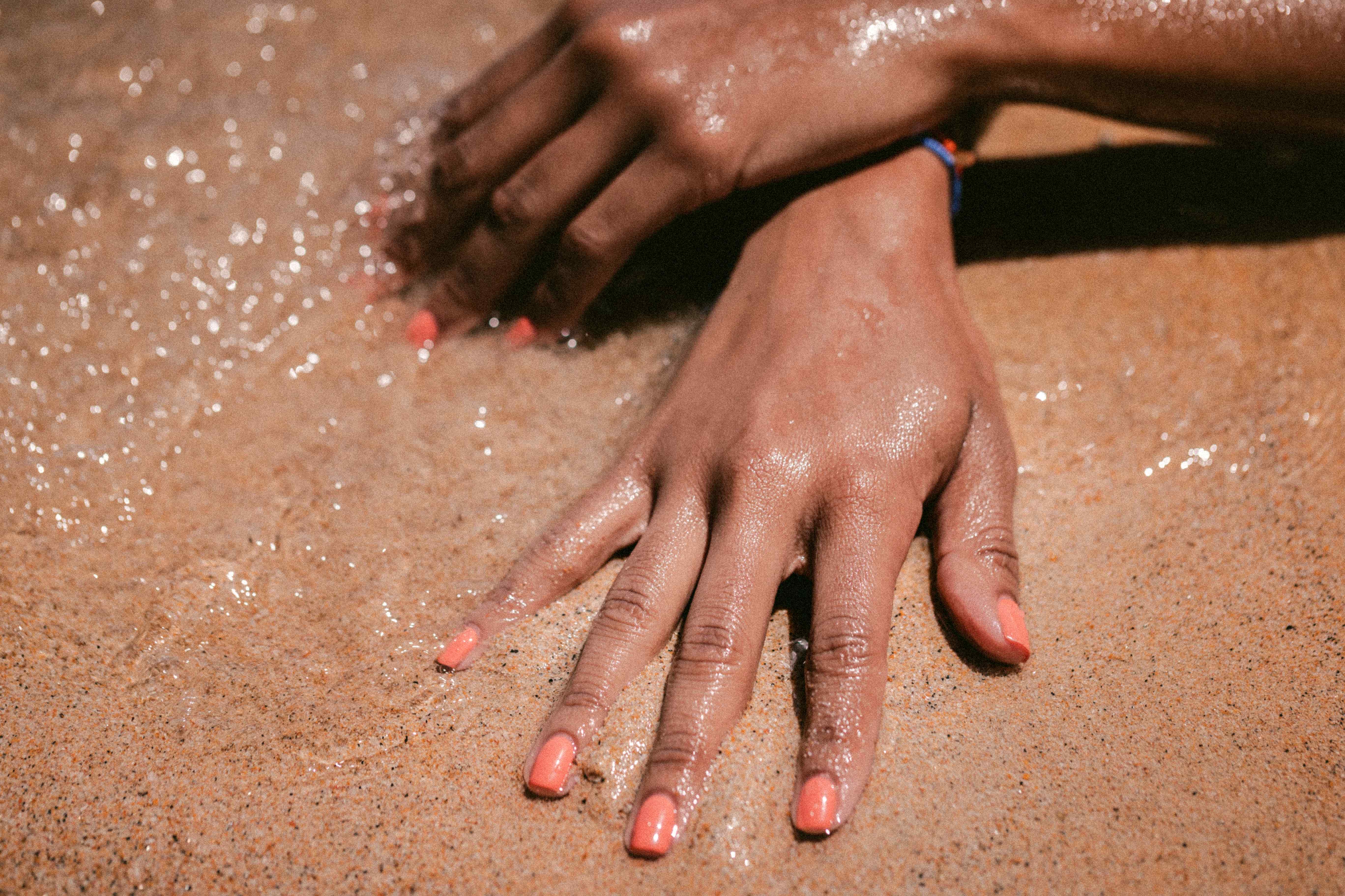 Körper – Frau mit orange lackierten Fingernägeln legt Hände am Ufer in den Sand