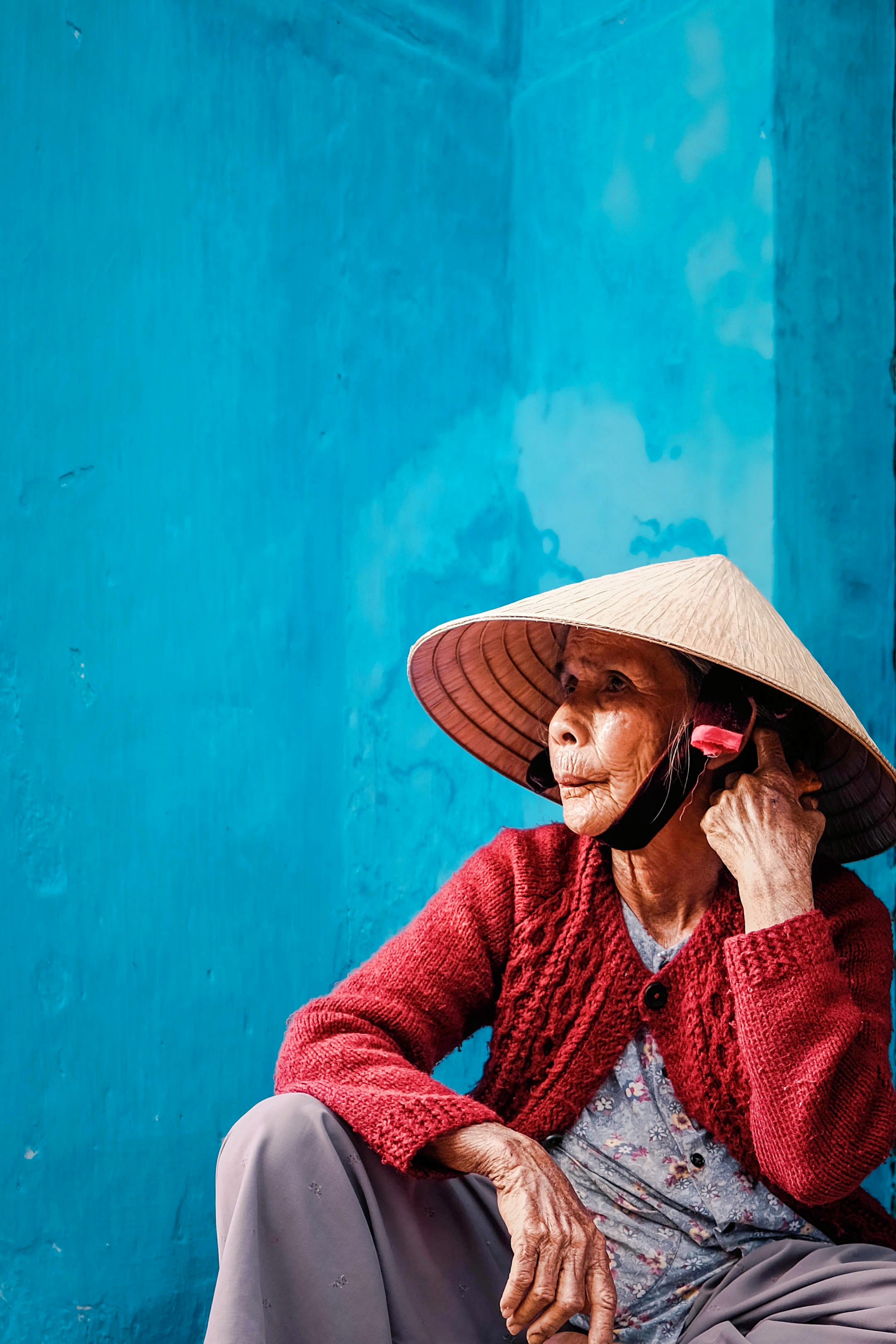 Ältere Frau mit Basthut, roter Strickjacke vor türkiser Wand