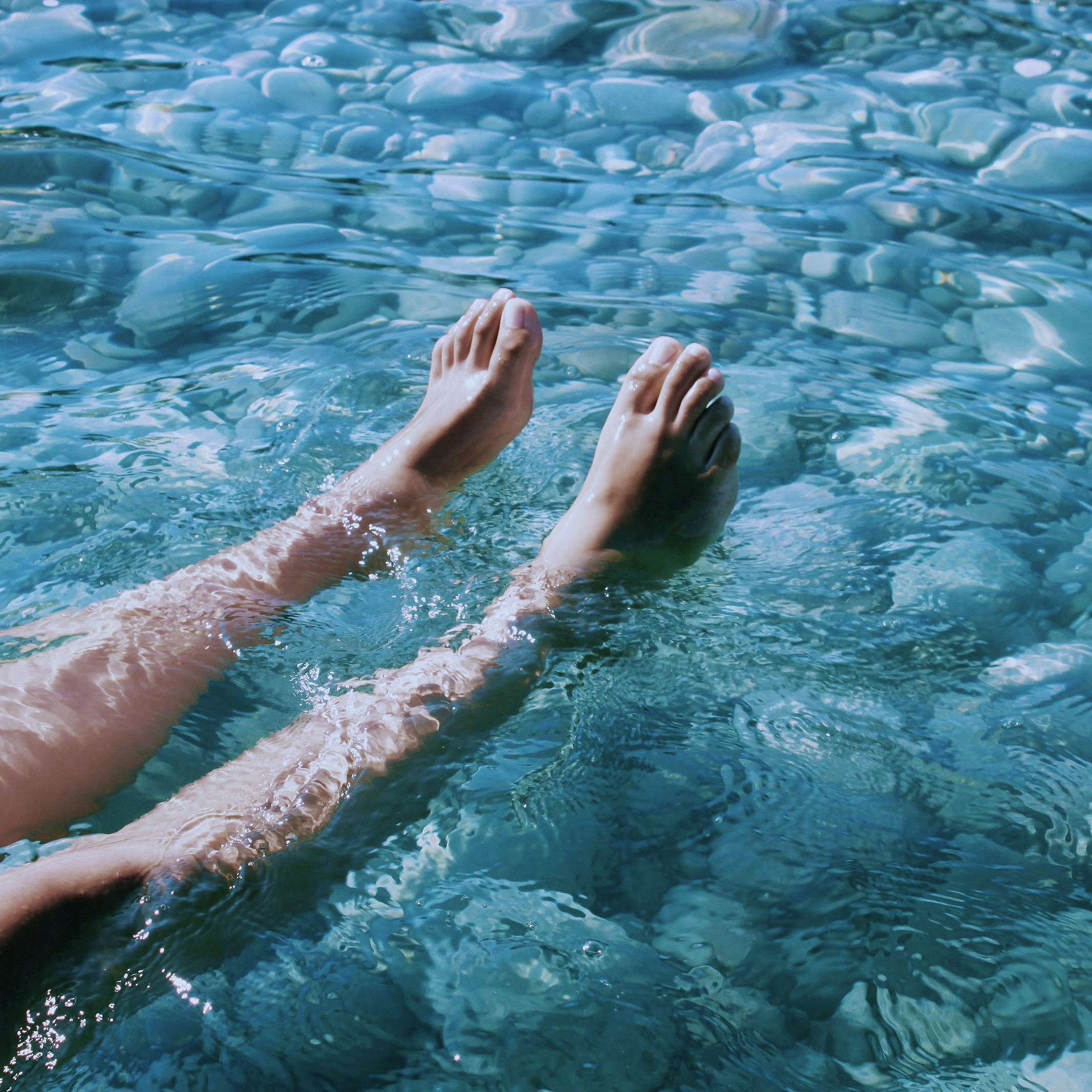 Nackte Beine und Füße in transparent-blauem Wasser über steinigem Grund